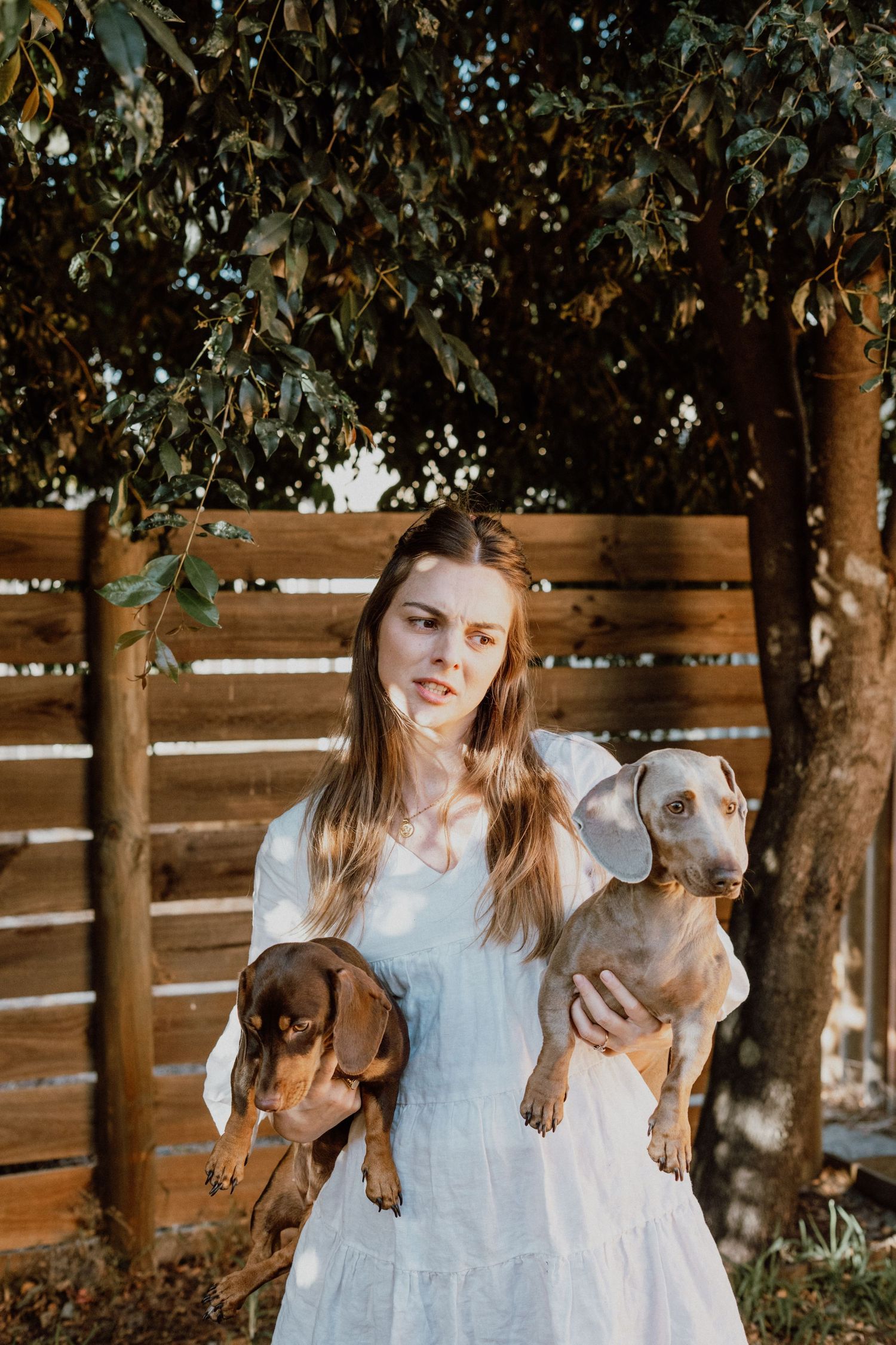 Young woman in white dress holding two dachshund dogs outdoors near wooden fence and green trees.