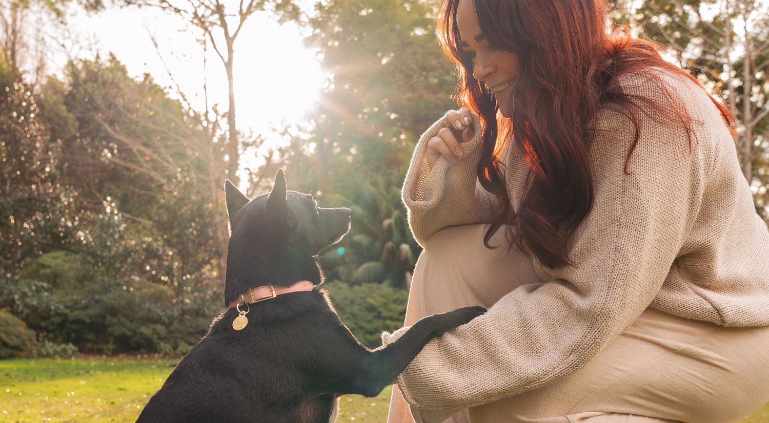 Red-haired woman kneeling in a sunlit garden petting a black dog wearing a collar.