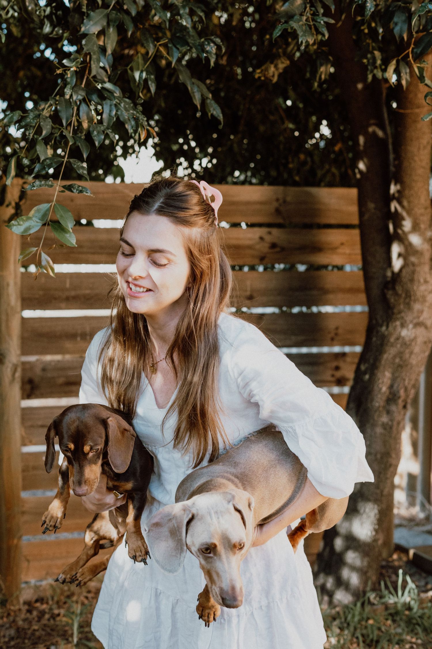 Young woman in white dress smiling while holding two dachshund dogs outdoors near wooden fence.