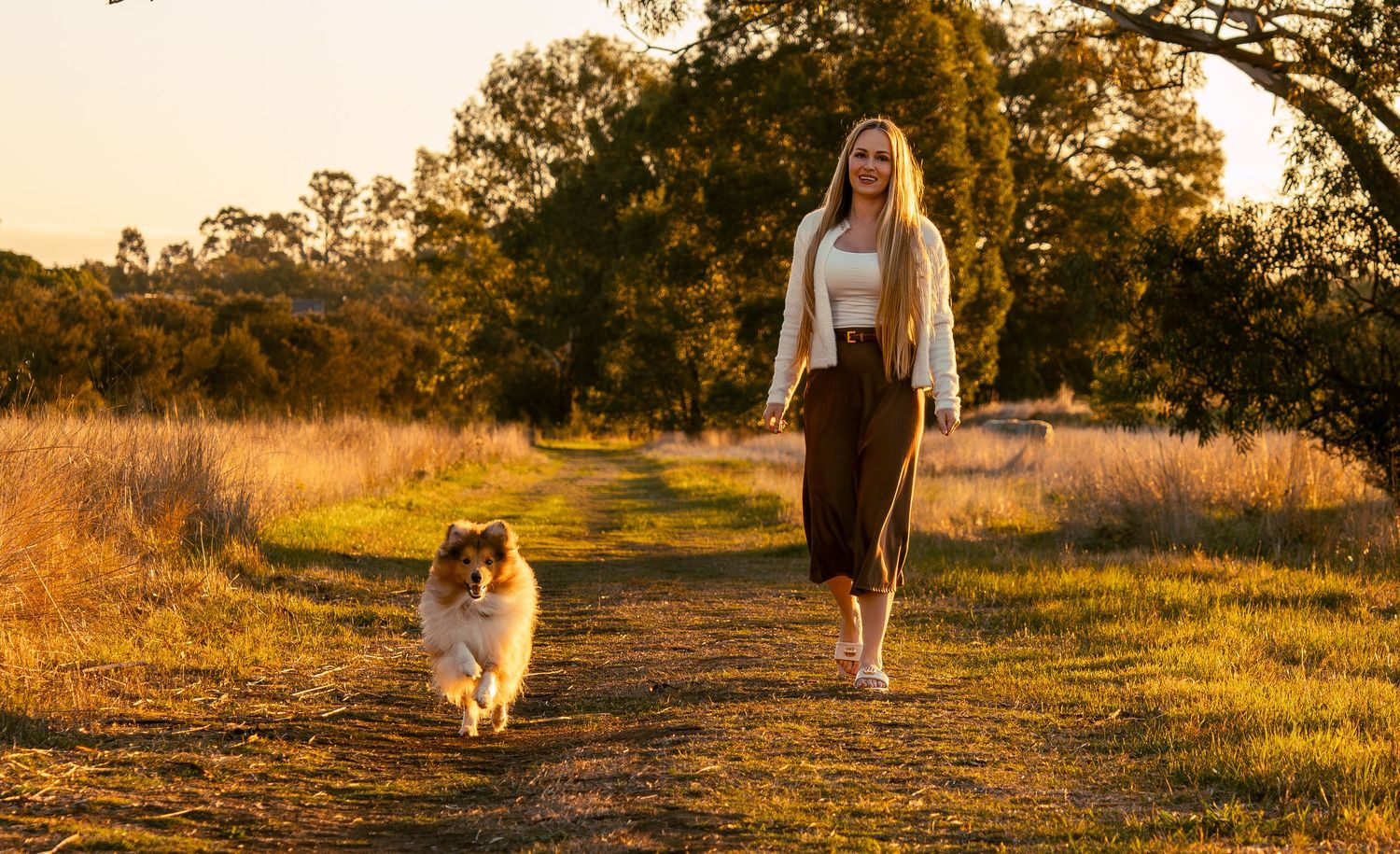 Smiling young woman walks with her Rough Collie dog along a dirt path in warm sunset light.