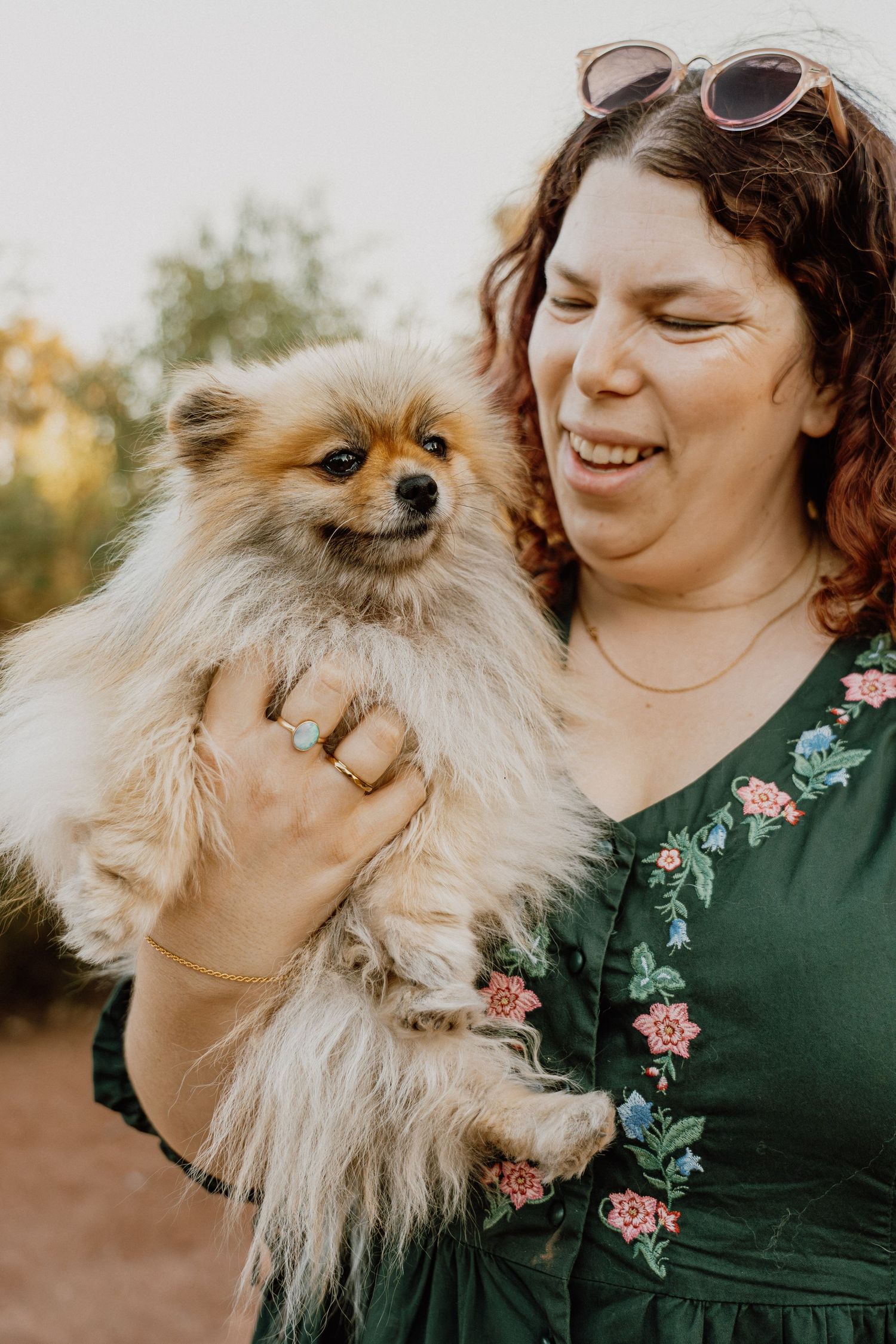 Smiling woman in green floral dress holds a fluffy orange Pomeranian dog outdoors at dusk.