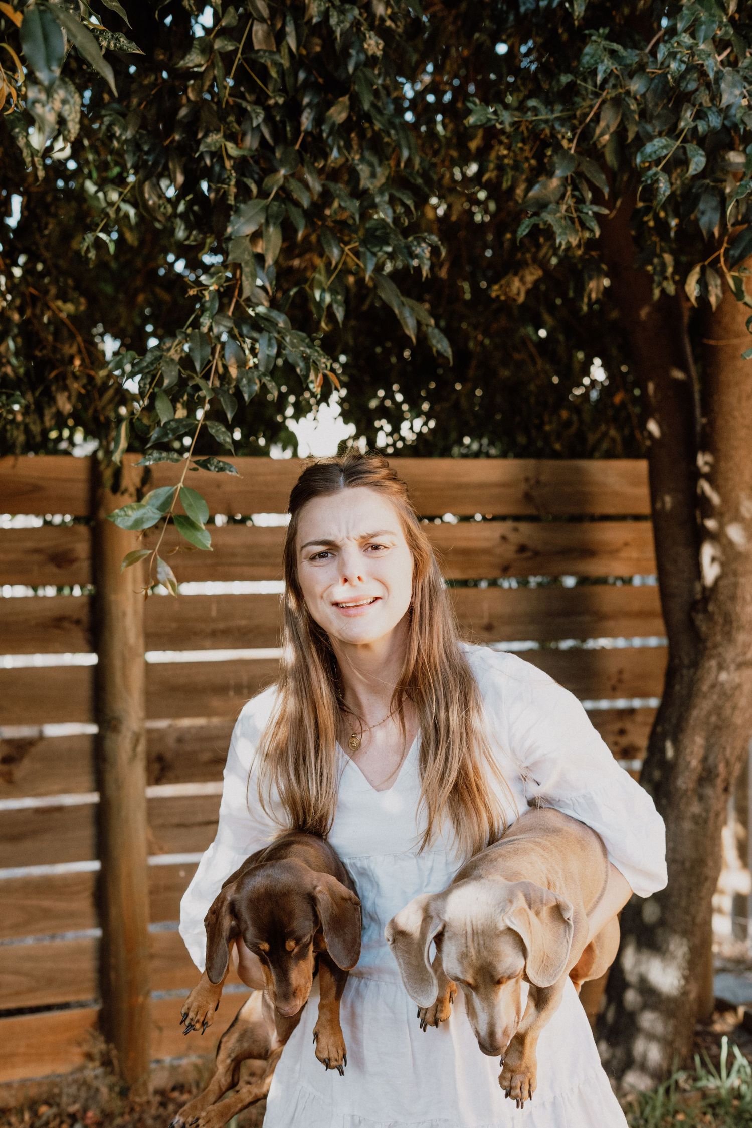 Woman in white dress holding two dachshund dogs outdoors near wooden fence and green trees.