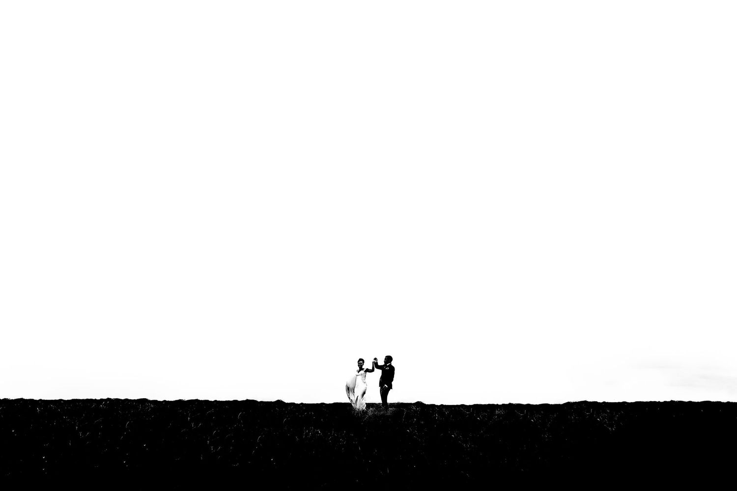 Black and white photo of a couple standing together on a hilltop, silhouetted against a bright white sky.