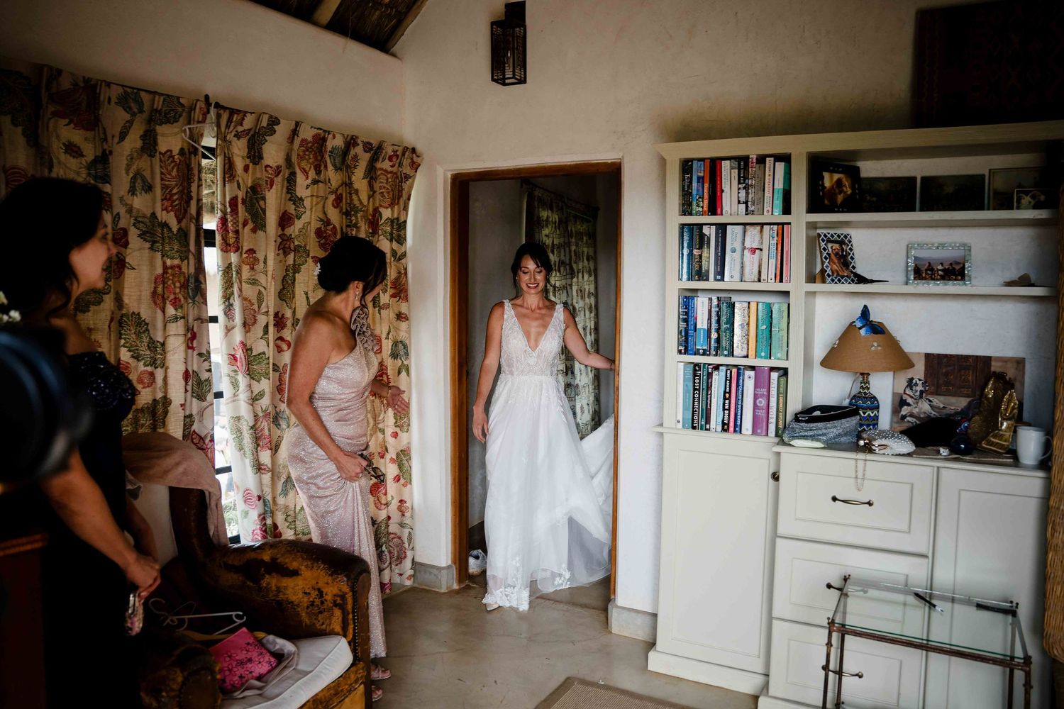 A white wedding dress is displayed in a cozy room with floral curtains and built-in bookshelves.