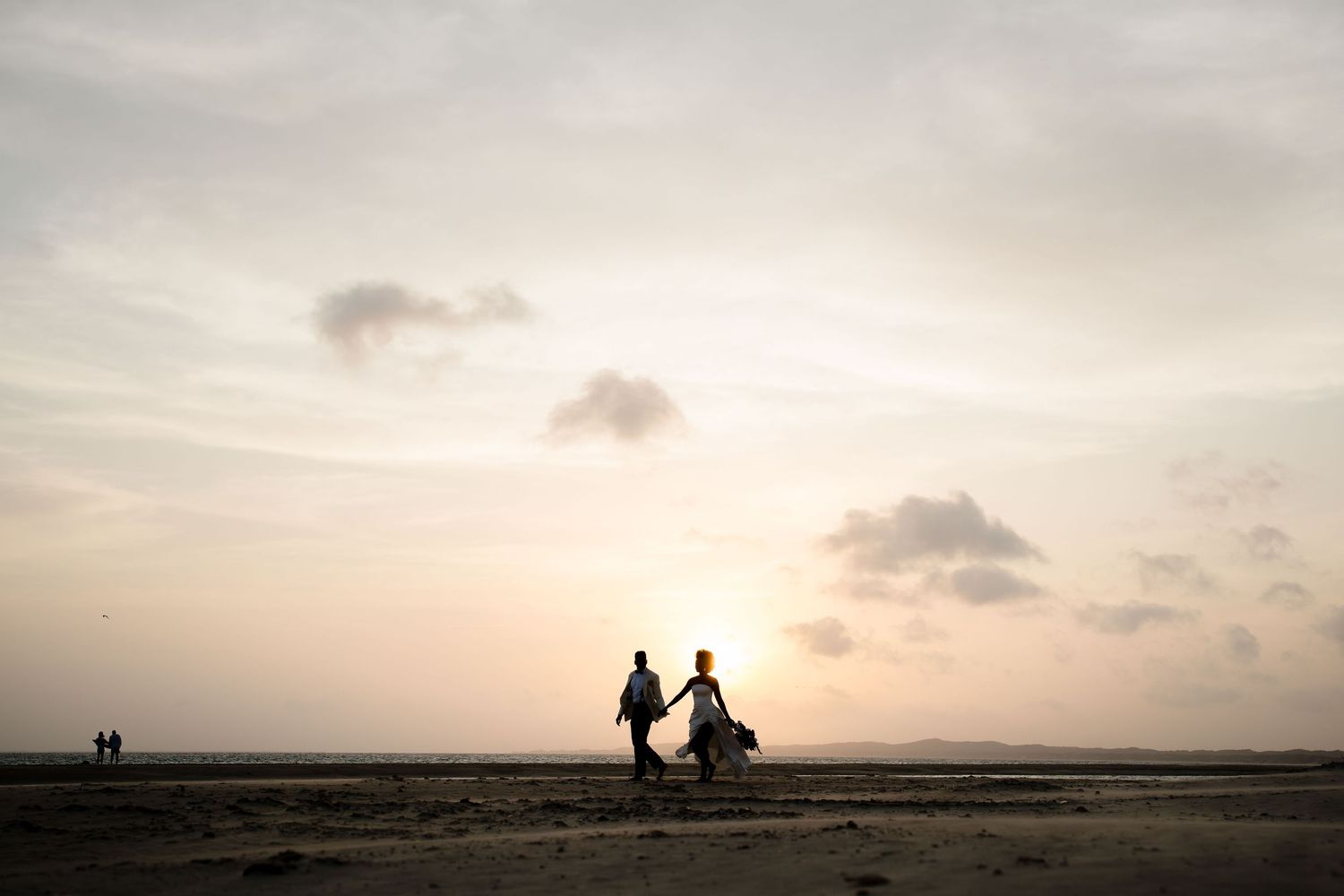 Silhouettes of two people walking on a beach at sunset with soft clouds in a warm sky.