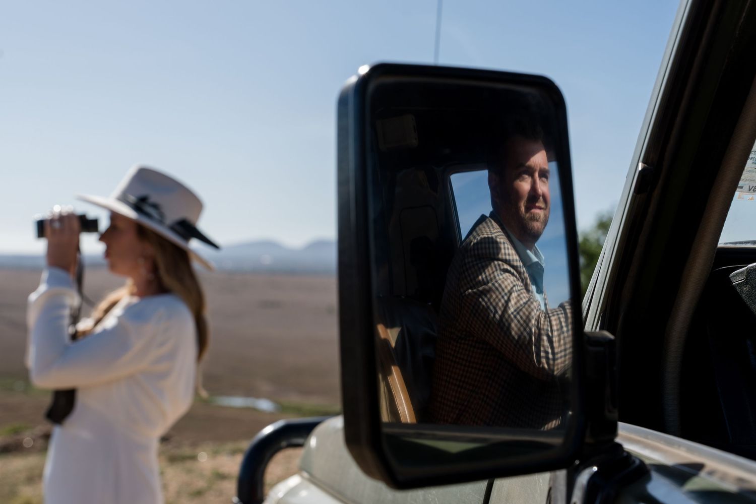 A safari scene with someone using binoculars while another person is reflected in the vehicle mirror.