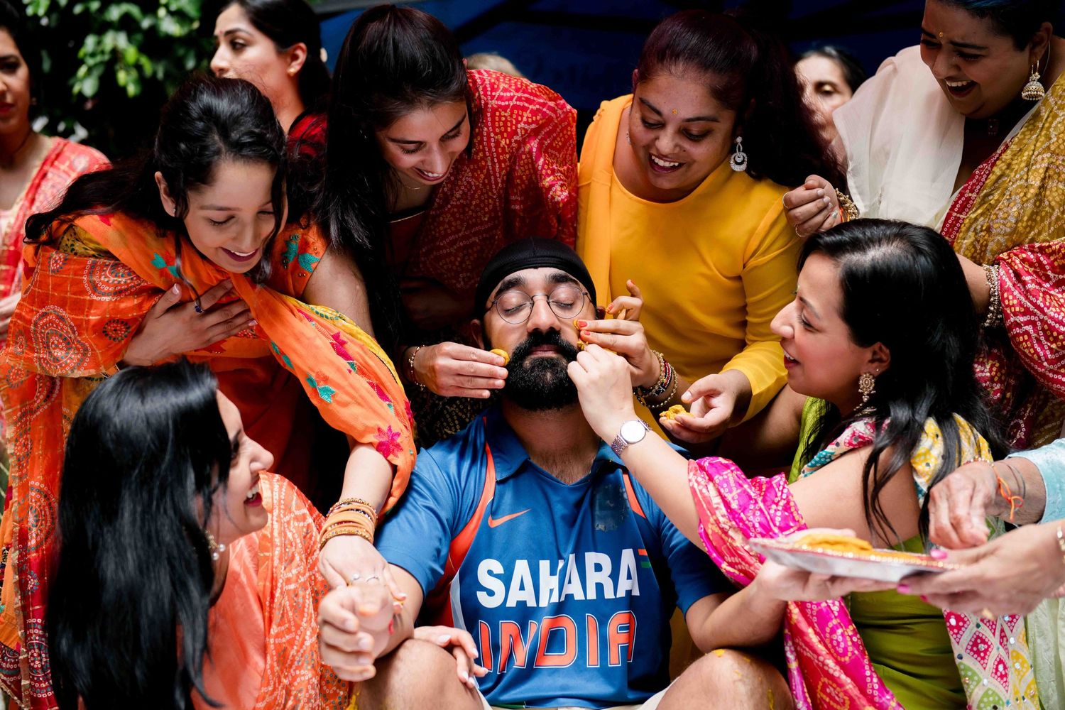 Group playfully feeding sweets during festive Indian pre-wedding celebration.