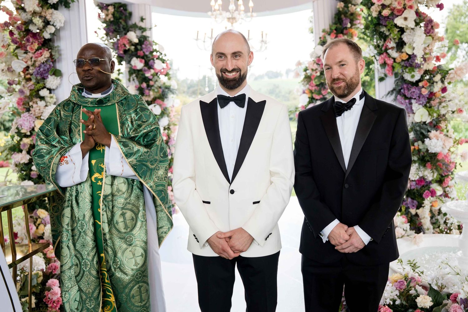 Wedding party members in black and white tuxedos and green attire pose together with floral decorations.