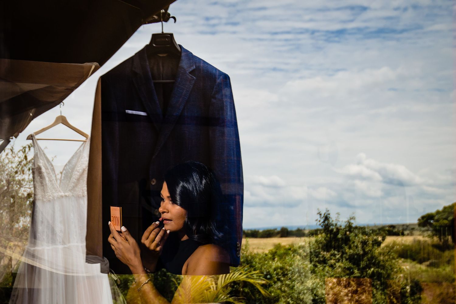 Wedding dress and suit hanging against a scenic outdoor landscape with cloudy sky.