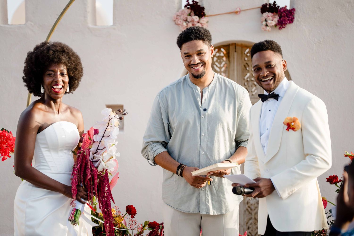 Three people smile together at a wedding celebration with red floral decorations and a white backdrop.