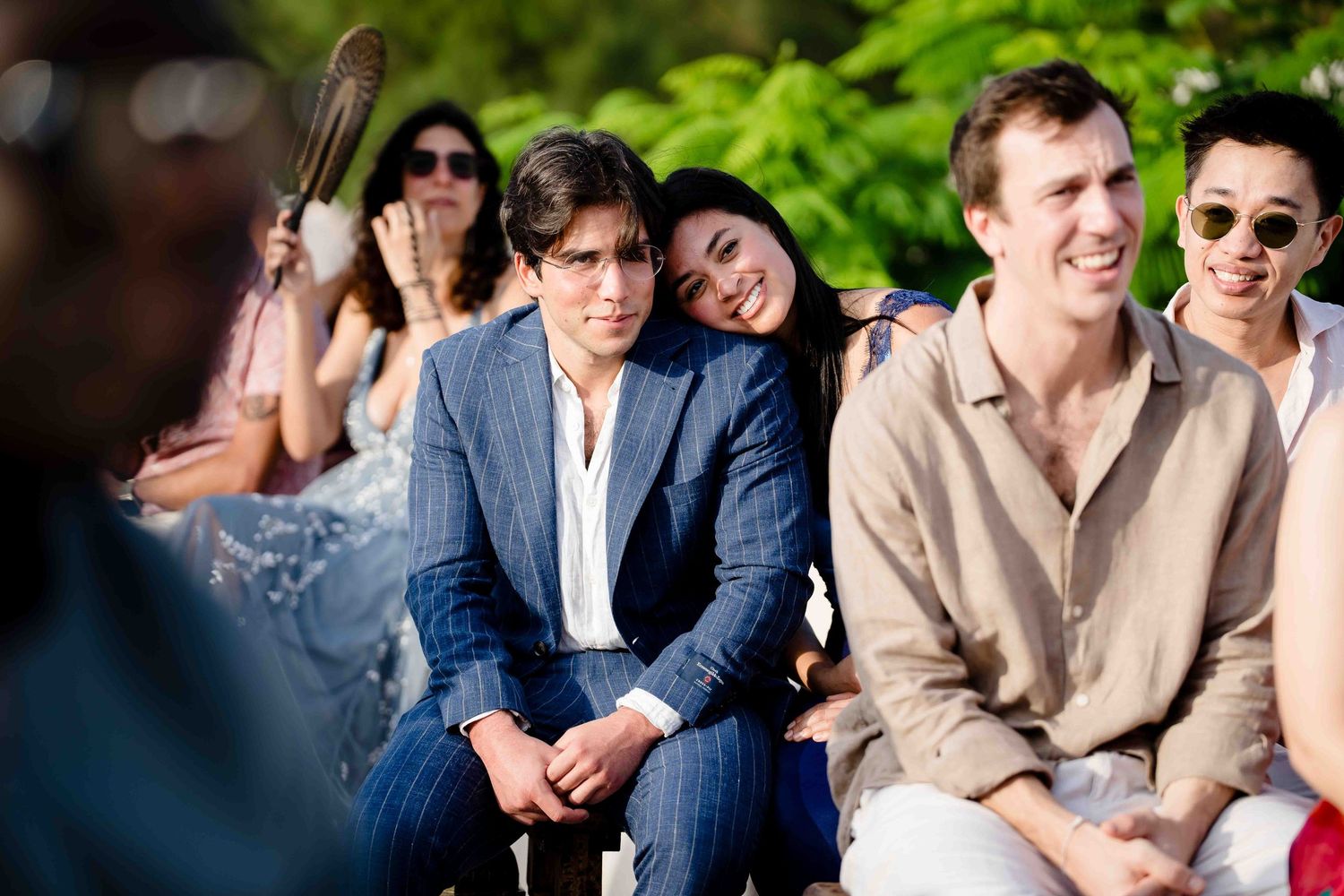 A group of people in formal attire laugh together outdoors during a sunny celebration.