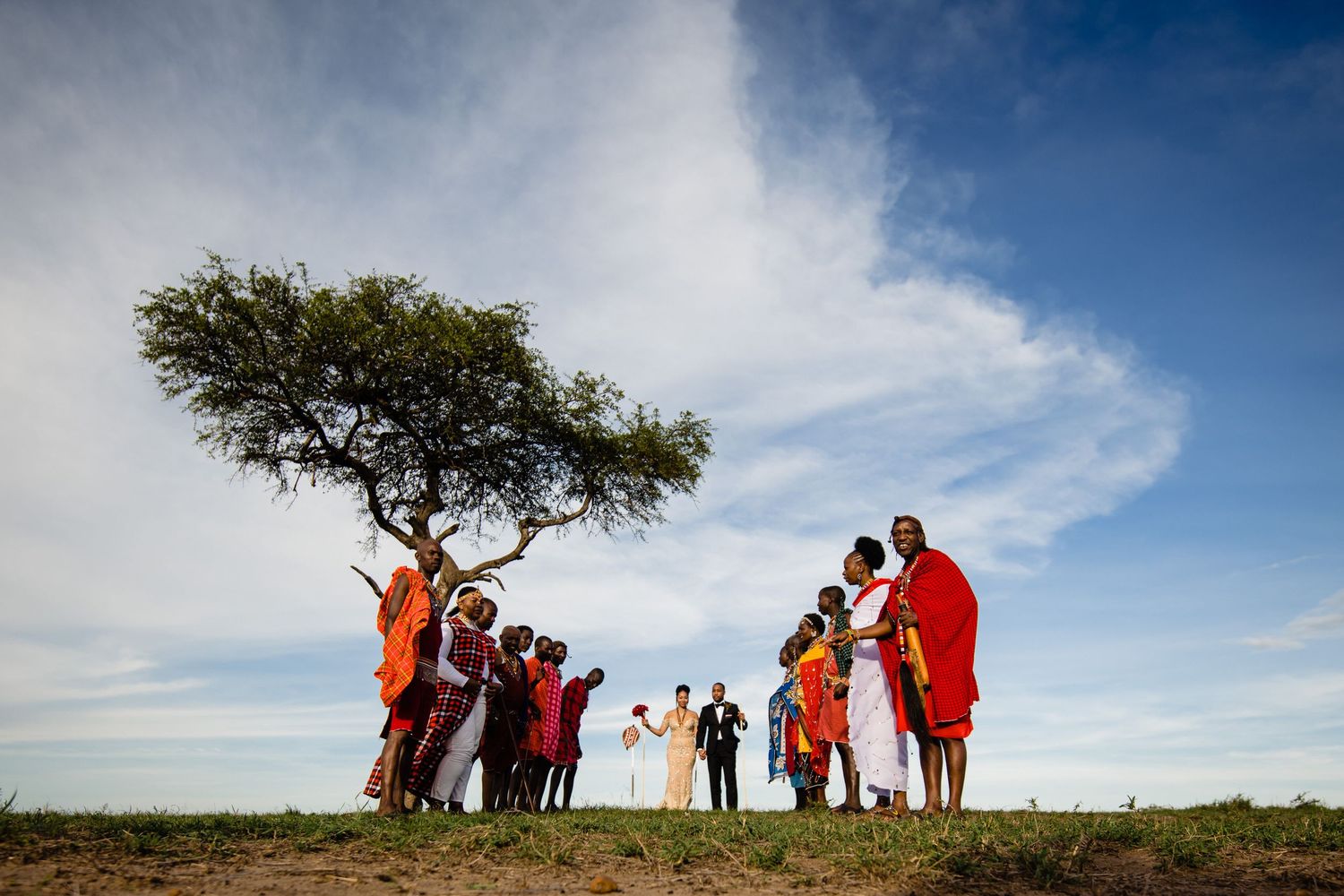 Silhouettes of people in traditional African dress gathered under an acacia tree against a dramatic blue sky.