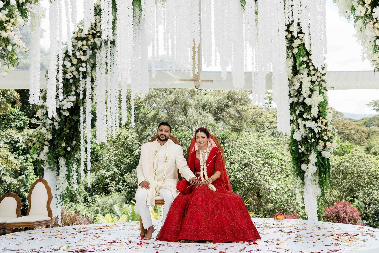 A wedding couple sits under a white draped canopy with green vines and flowers at an outdoor ceremony.