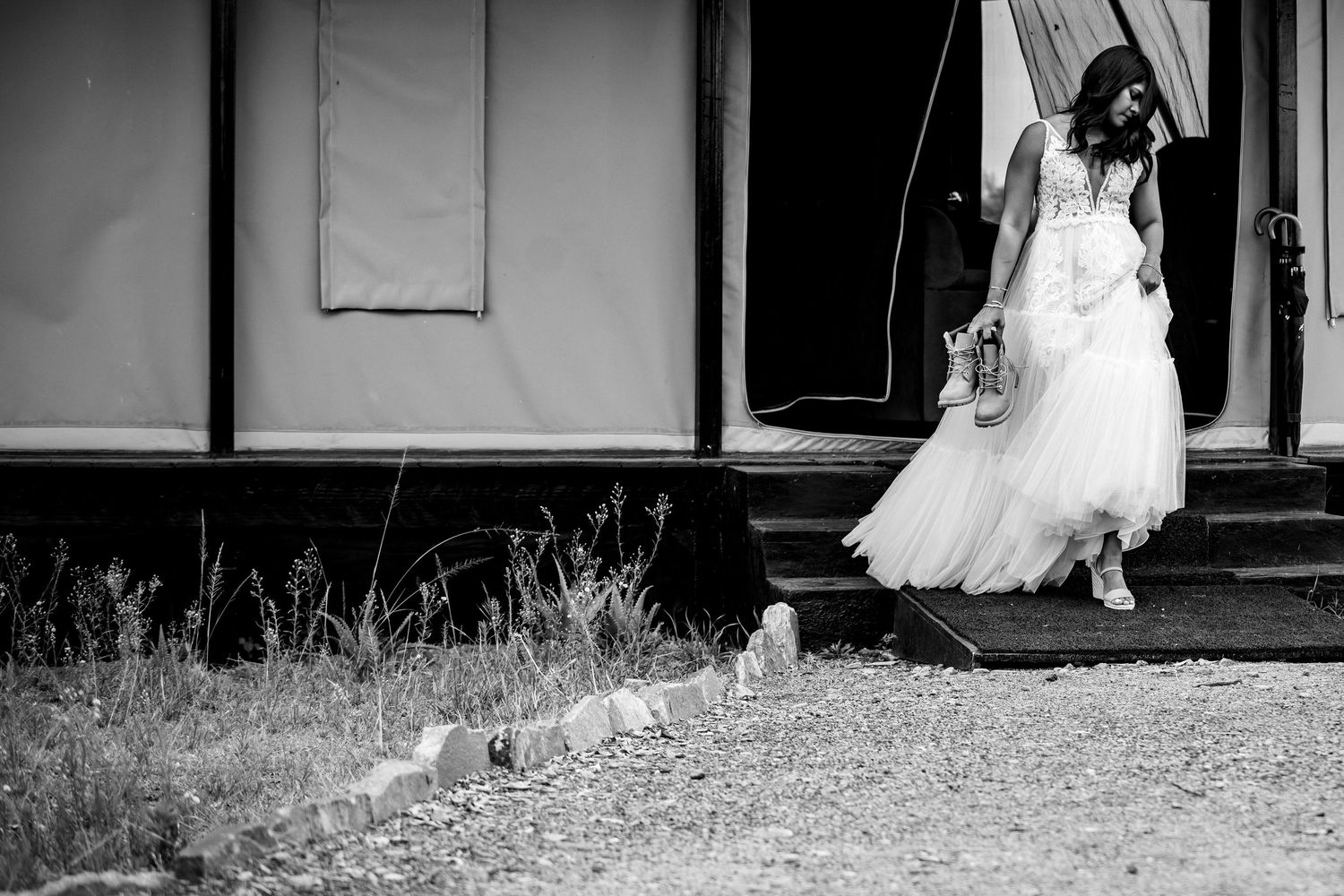 Black and white photo of a bride in a white gown standing outside a tent venue, holding shoes.