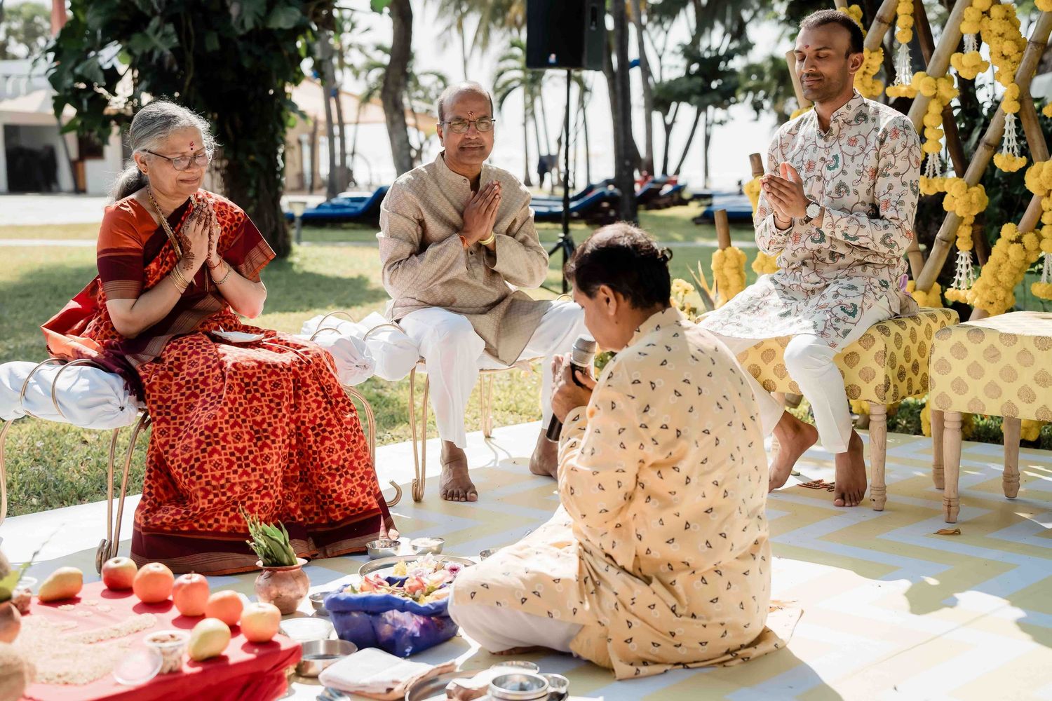 People sit together outdoors participating in a traditional Thai ceremony with offerings and decorations arranged around them.