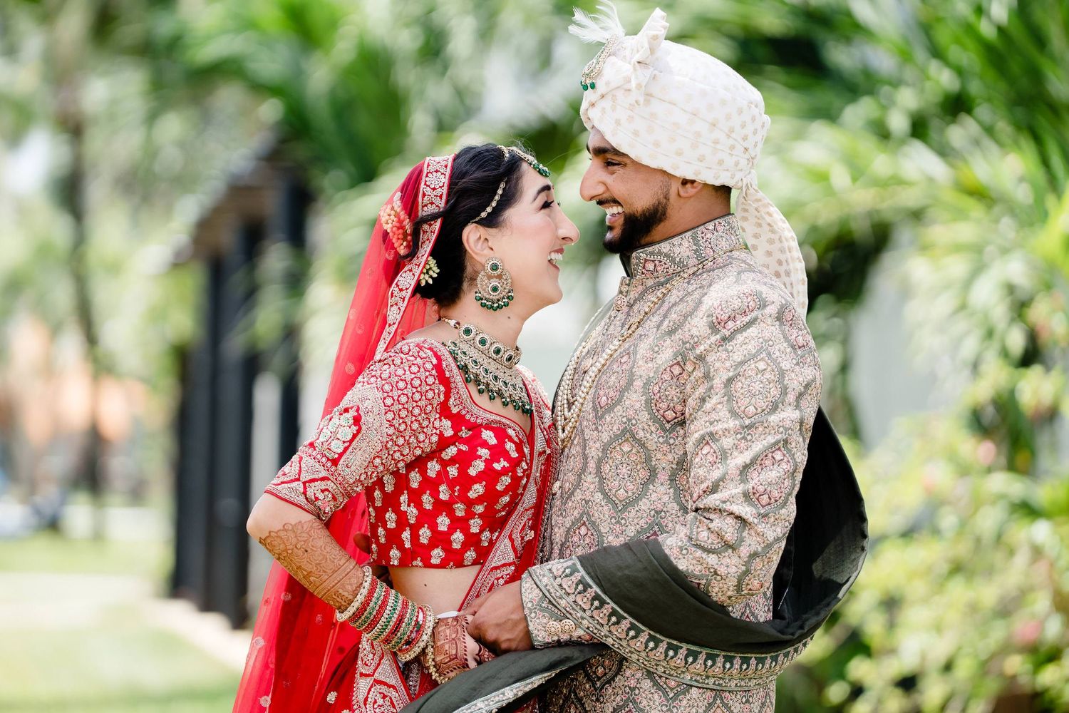 A couple in traditional Indian wedding attire embraces during their outdoor celebration surrounded by greenery.
