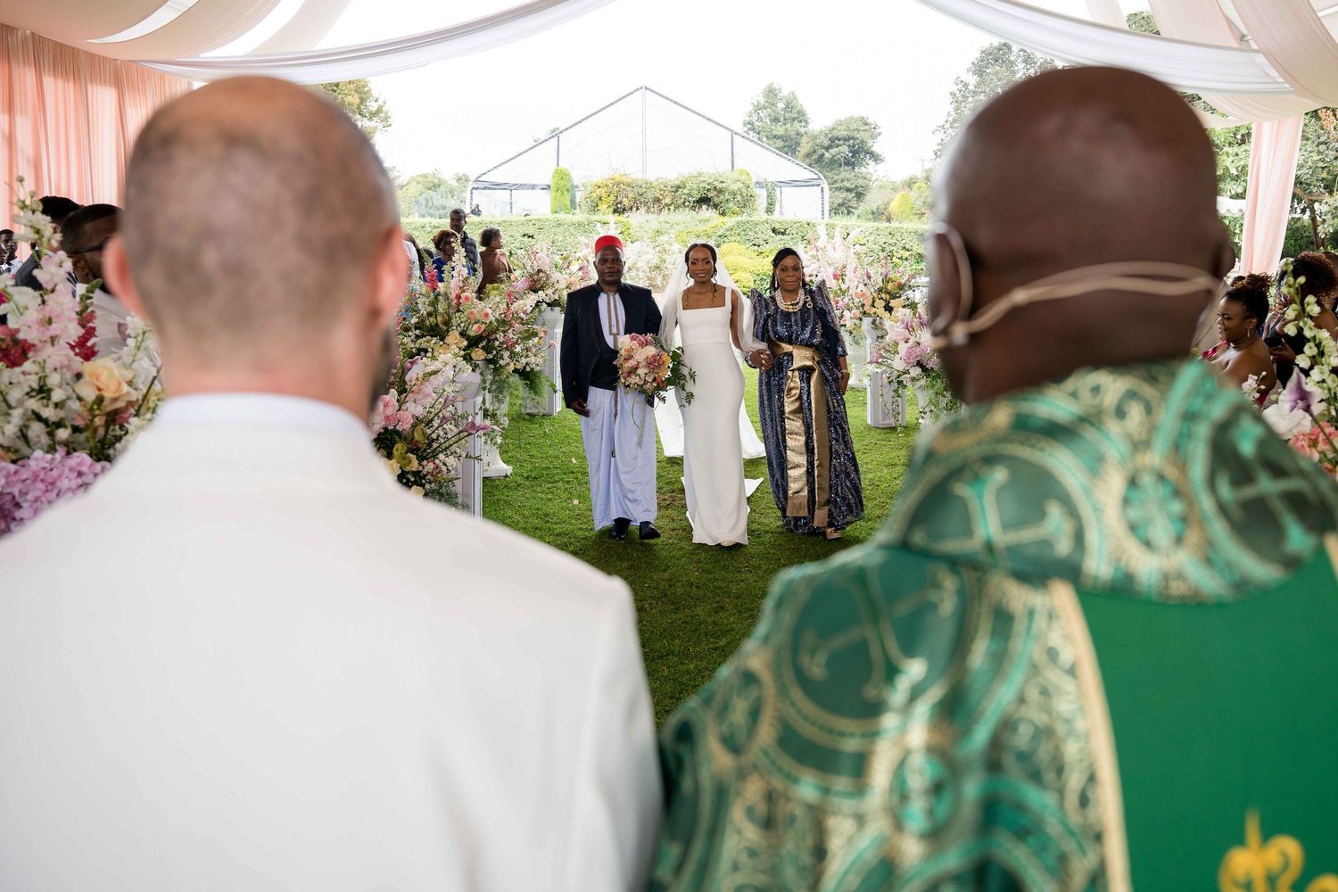 An outdoor wedding ceremony under a white tent with floral decorations and clergy in green vestments.