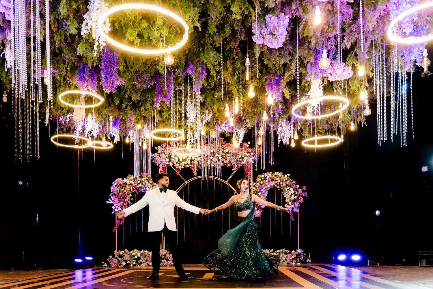 A couple dances beneath illuminated neon rings and floral arrangements in a stunning nighttime wedding reception setup.