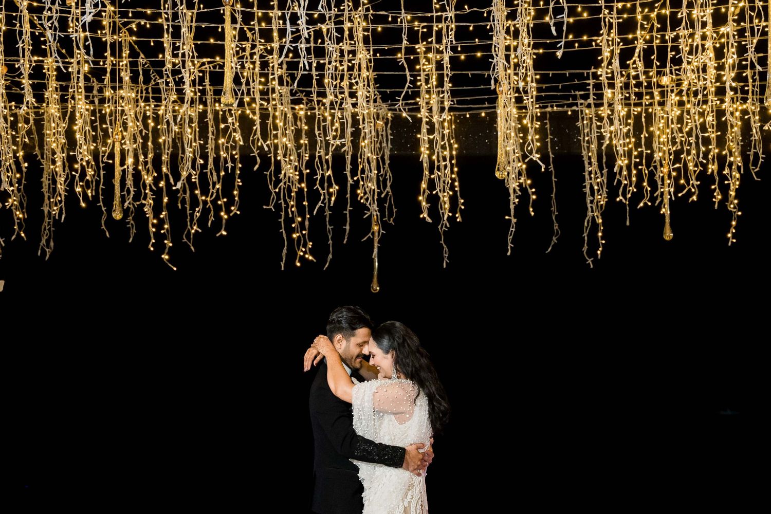 A couple dances together under a glowing canopy of warm string lights during a romantic evening celebration.