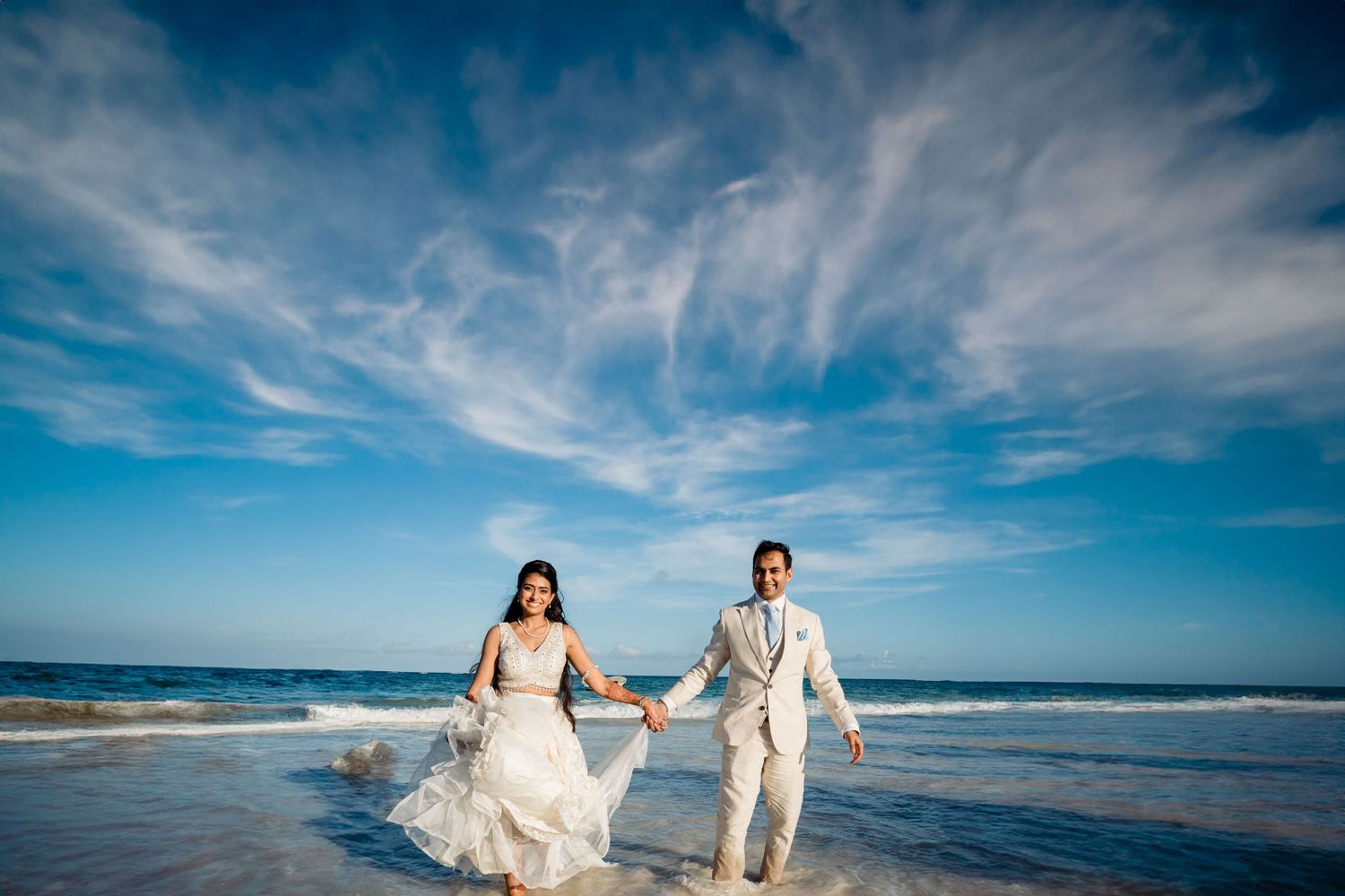 A couple walks hand in hand along the shoreline in formal attire.