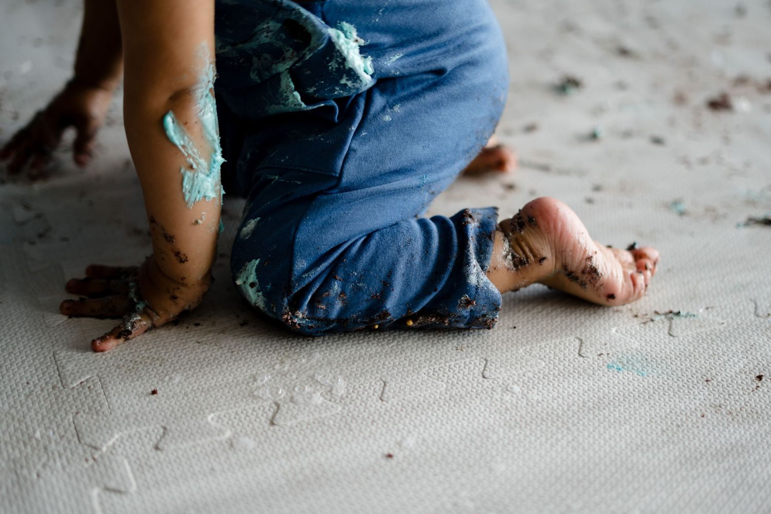 Small child in blue jeans kneels on a messy floor with turquoise paint and dirt scattered around.