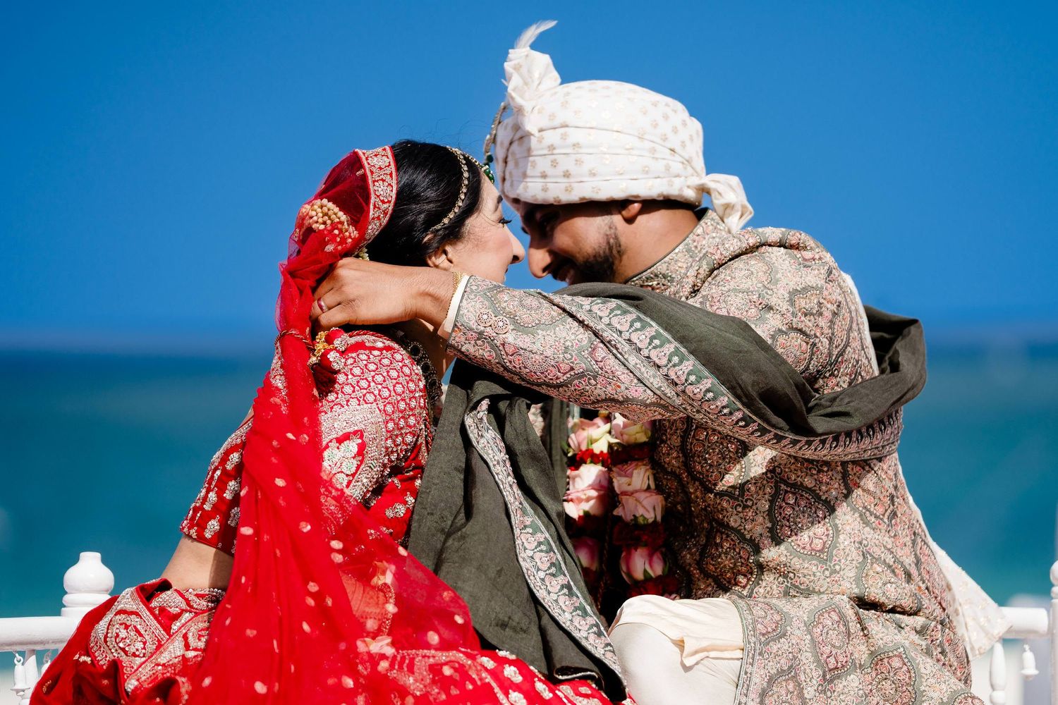A couple in traditional Indian wedding attire embraces on a sunny beach with turquoise water in the background.