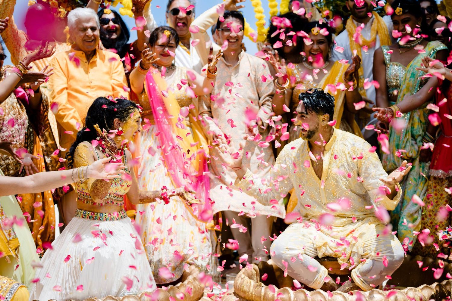 Colorful confetti and flower petals shower down on celebrating wedding party members dressed in light colored outfits.