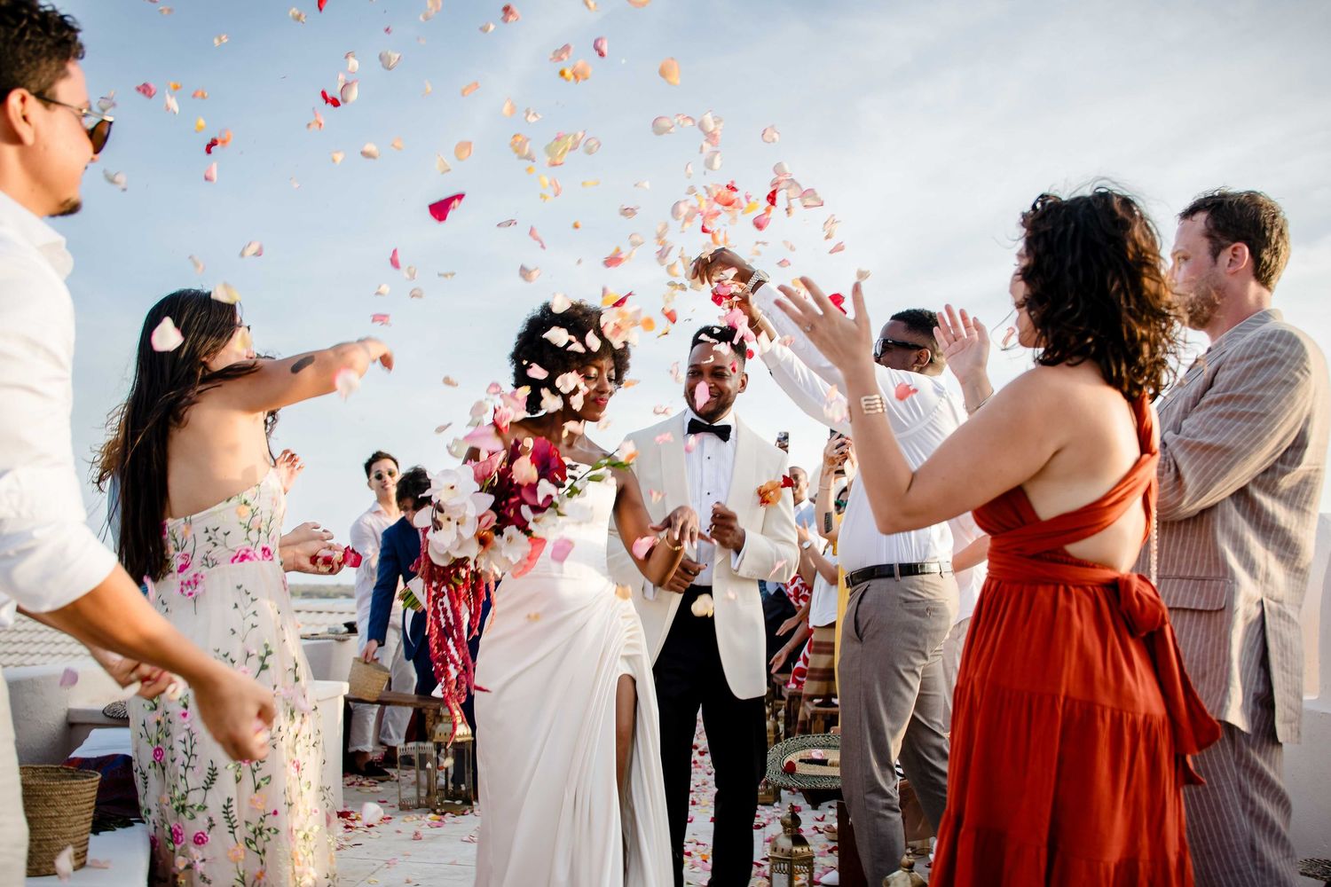 A joyful wedding celebration with guests throwing pink and red flower petals in the air at an outdoor venue.