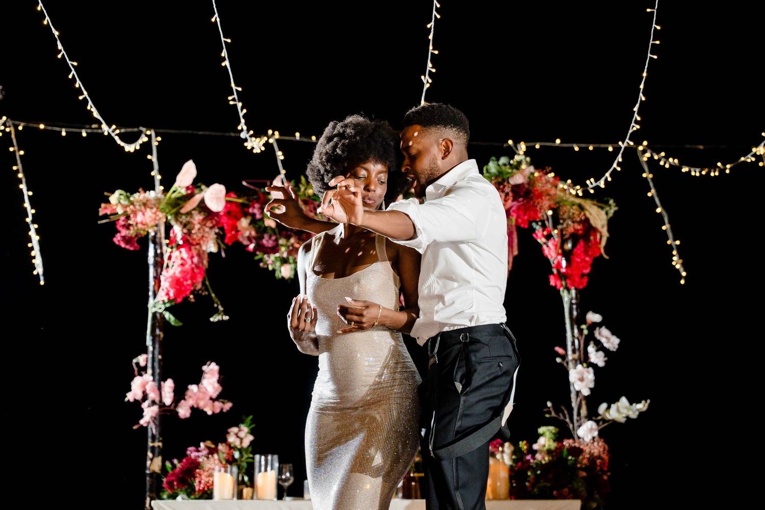 A couple shares a romantic first dance at their wedding reception under twinkling string lights and floral decorations.