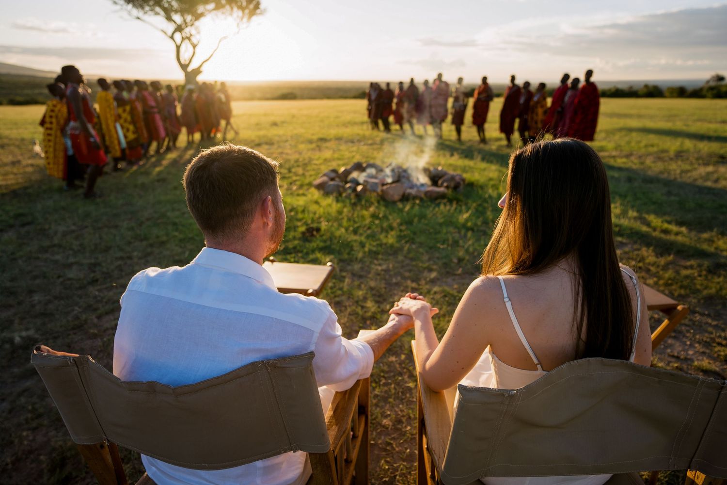 A couple sits in chairs watching a traditional ceremony with tribal dancers at sunset.