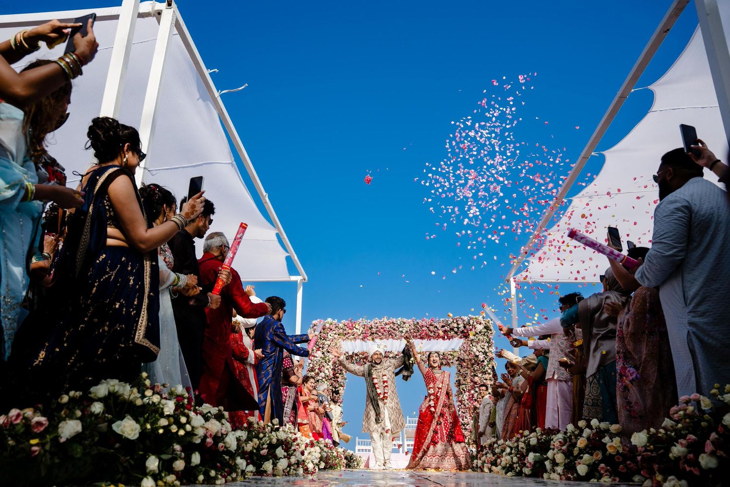 Wedding guests throw confetti and celebrate during an outdoor ceremony under bright blue skies.
