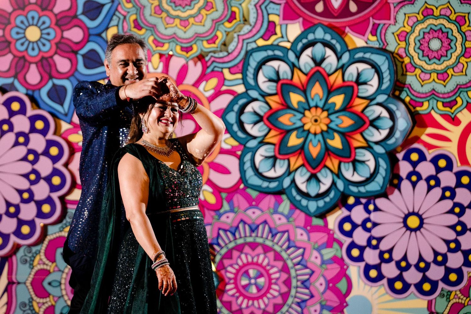 A couple poses in formal attire against a vibrant wall featuring colorful mandala patterns and floral designs.