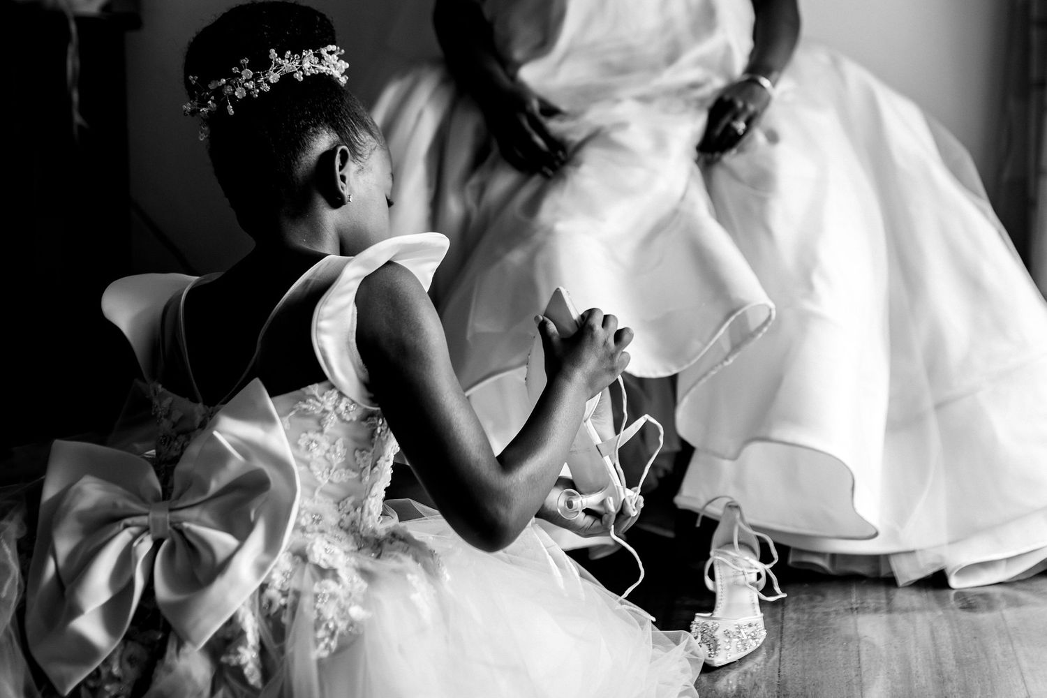 Black and white image of a small flower girl in white dress sitting next to wedding gown.
