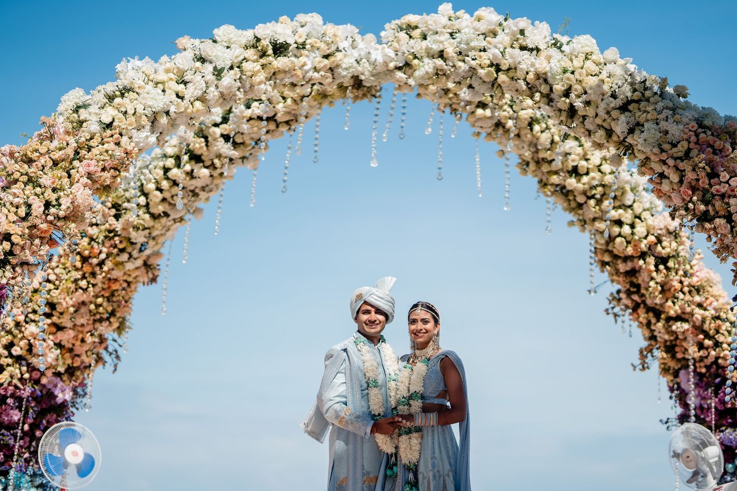 Couple posing under ornate floral arch during traditional Indian wedding ceremony.