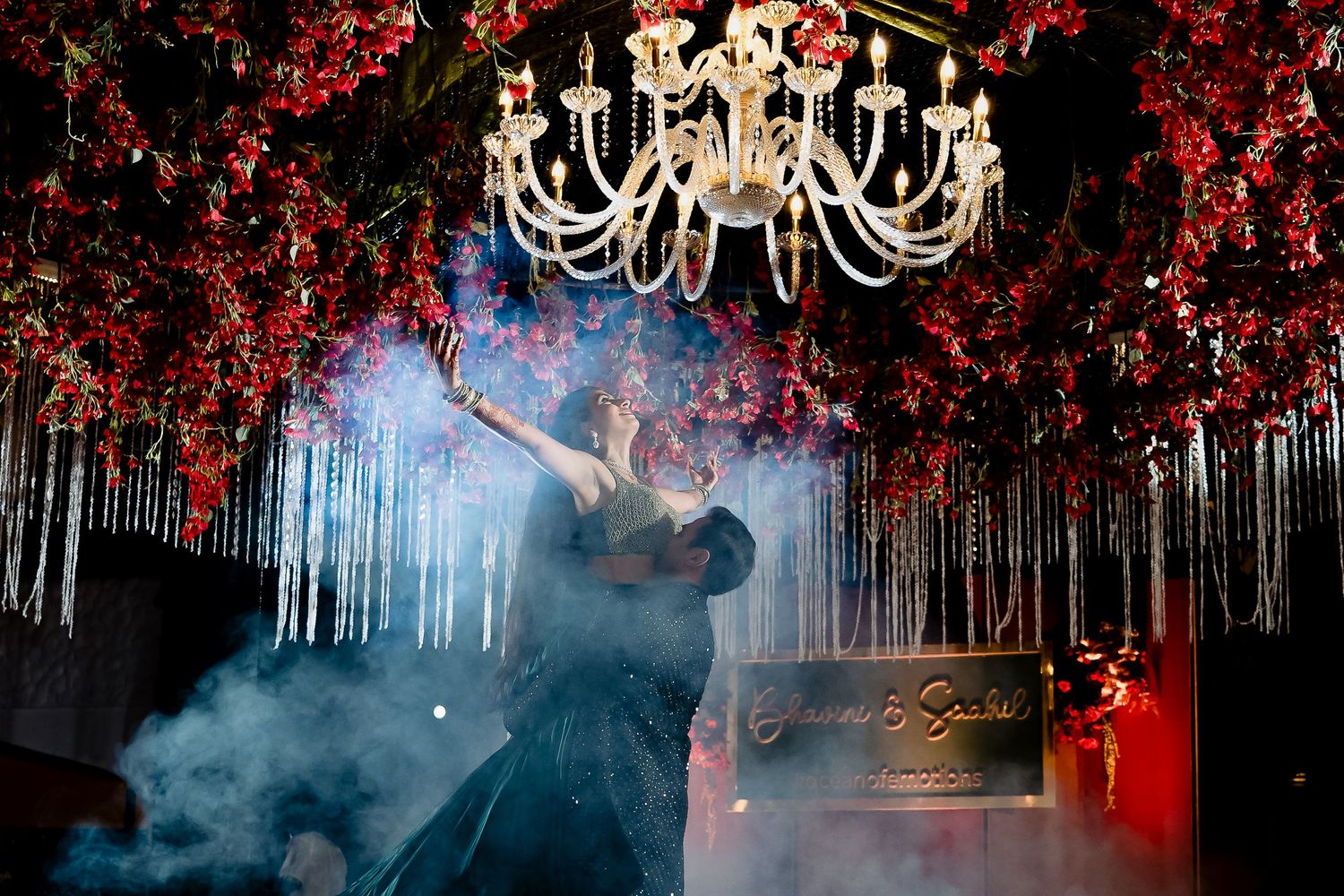Dramatic dance performance with fog effects under a red floral chandelier ceiling.
