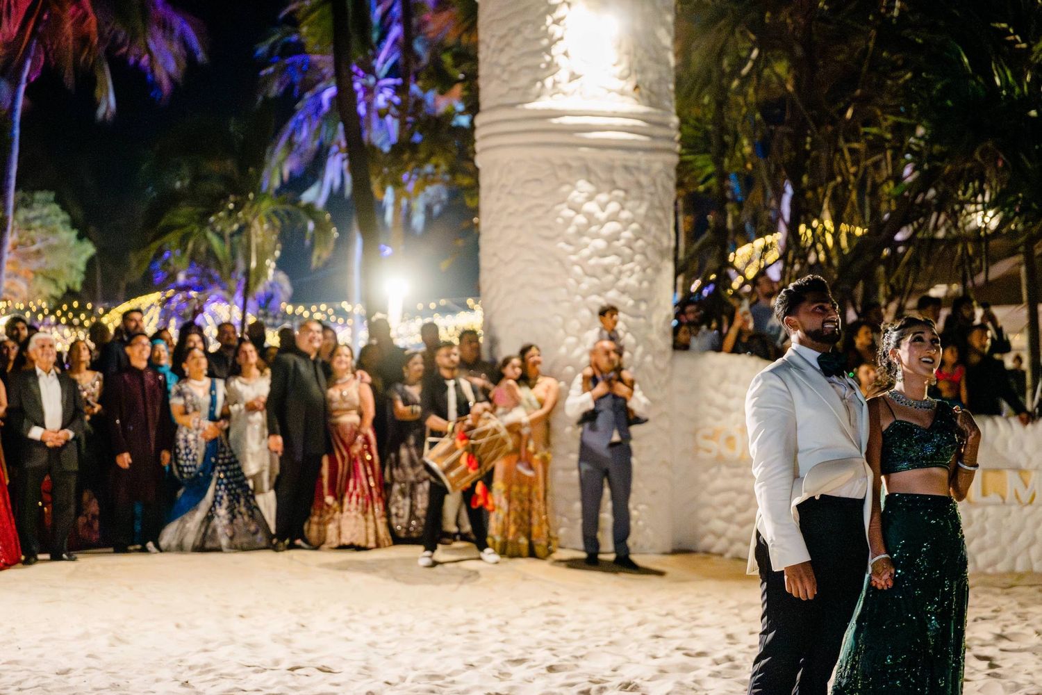 A festive nighttime beach party with guests gathered around while palm trees sway in the background.