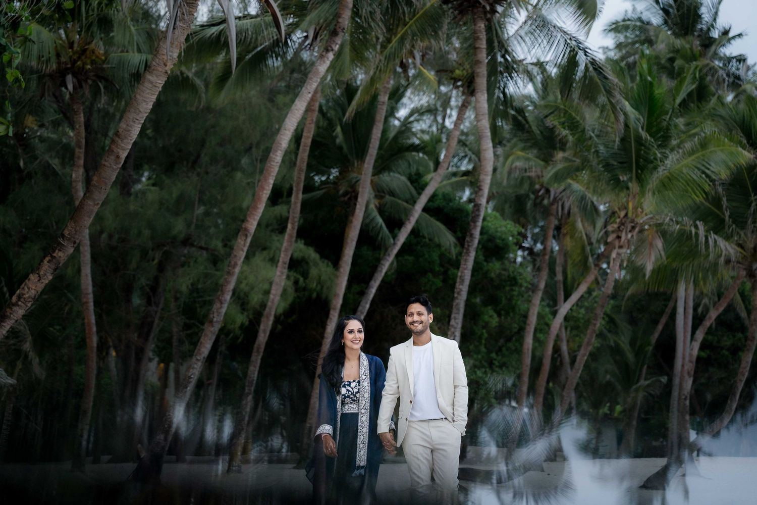 A couple walks together through a lush tropical palm grove with tall coconut trees surrounding them.