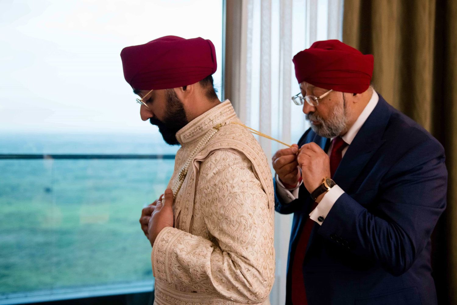 Two men in traditional Sikh turbans stand together near a window overlooking the ocean.