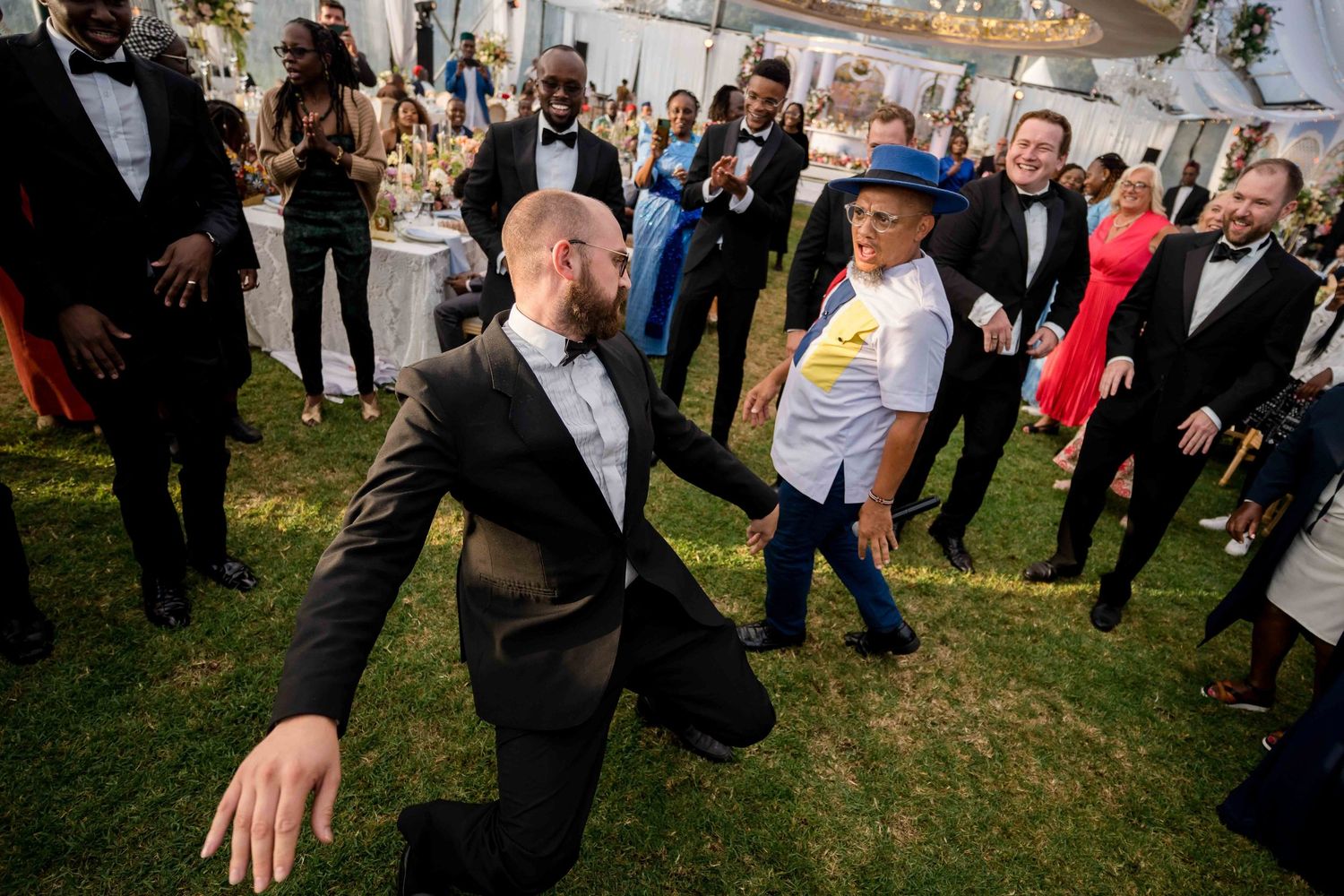 Wedding guests dance and celebrate together on a sunny outdoor lawn during a festive reception celebration.