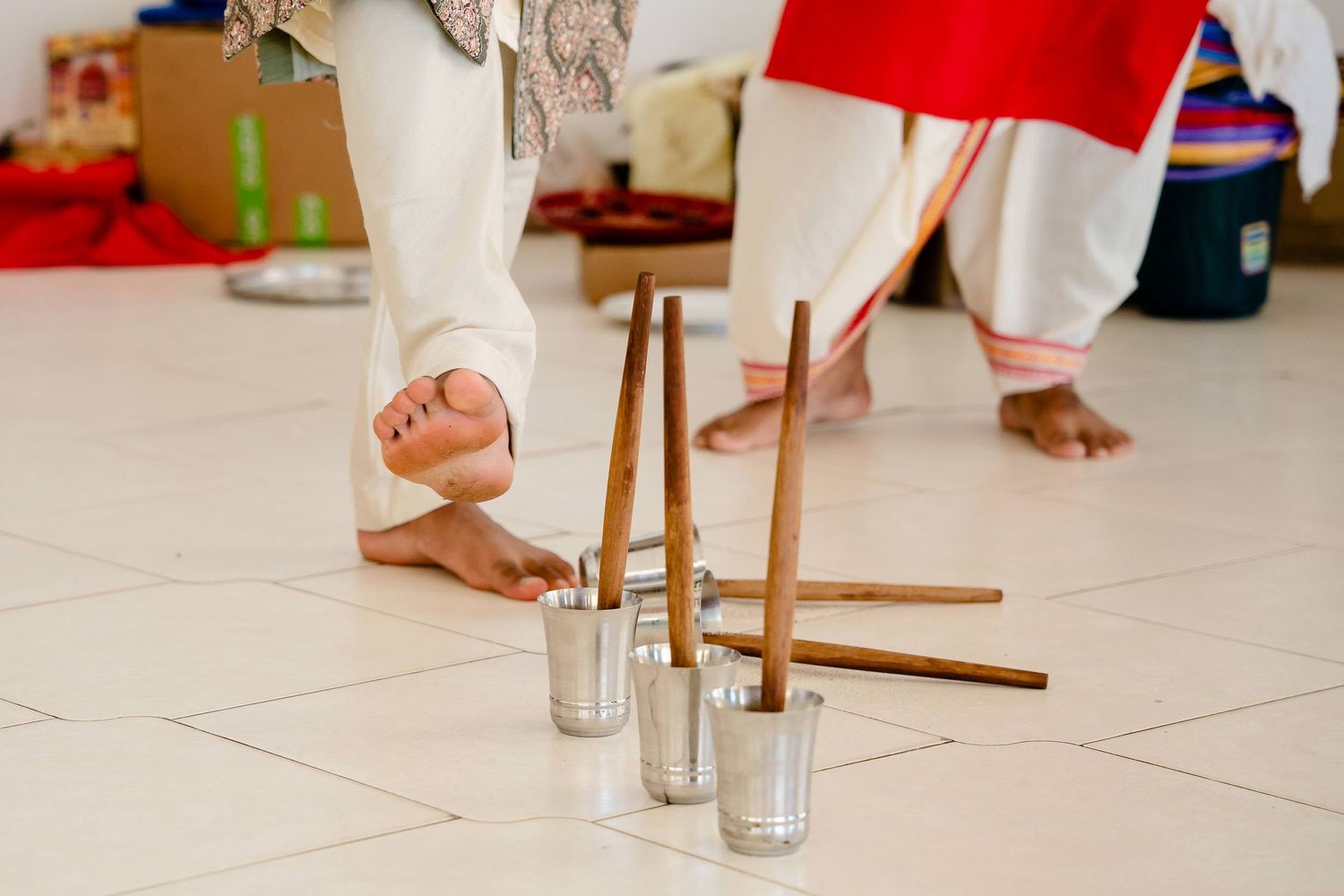 Bare feet stand near metal and wooden poles with small glass cups on white tile floor during a traditional ceremony.