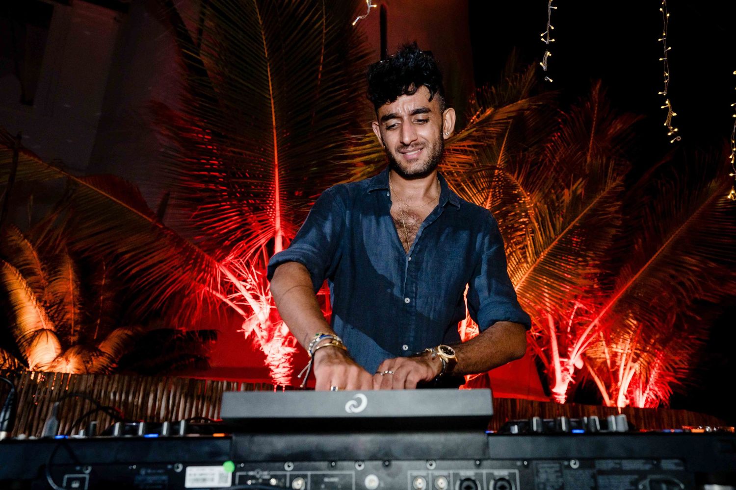 DJ performs at night with dramatic red lighting and palm trees creating an atmospheric outdoor party scene.