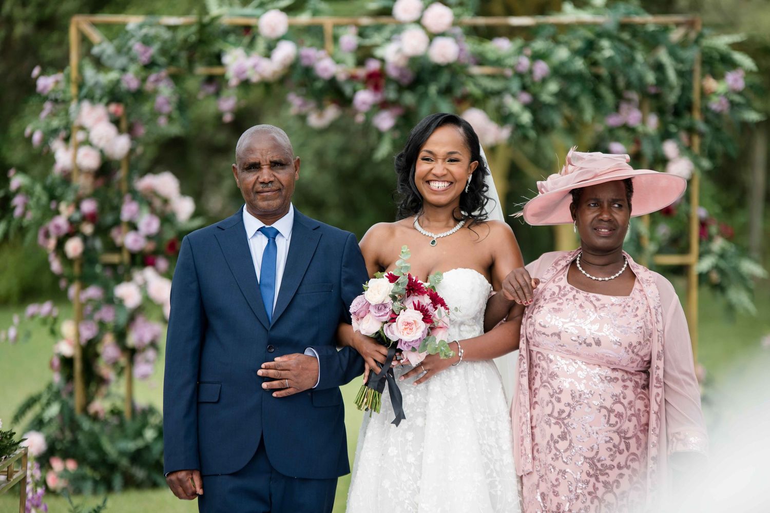 An elegant wedding party poses under a floral arch with pink and white blooms.