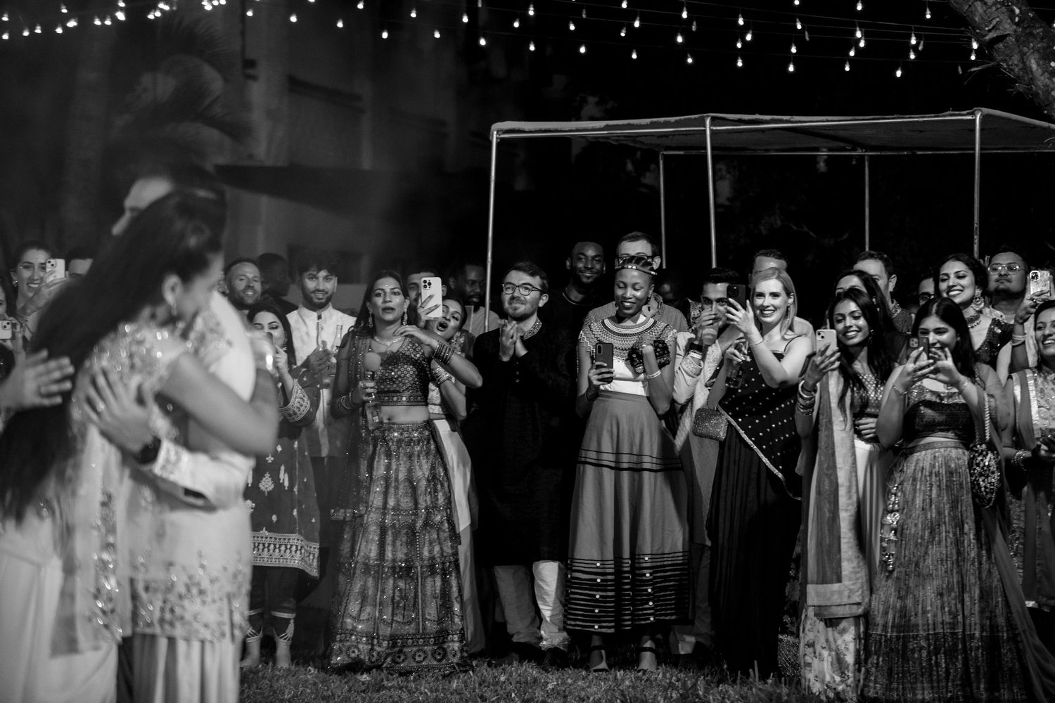 A black and white photo captures guests gathered under string lights at an outdoor celebration.