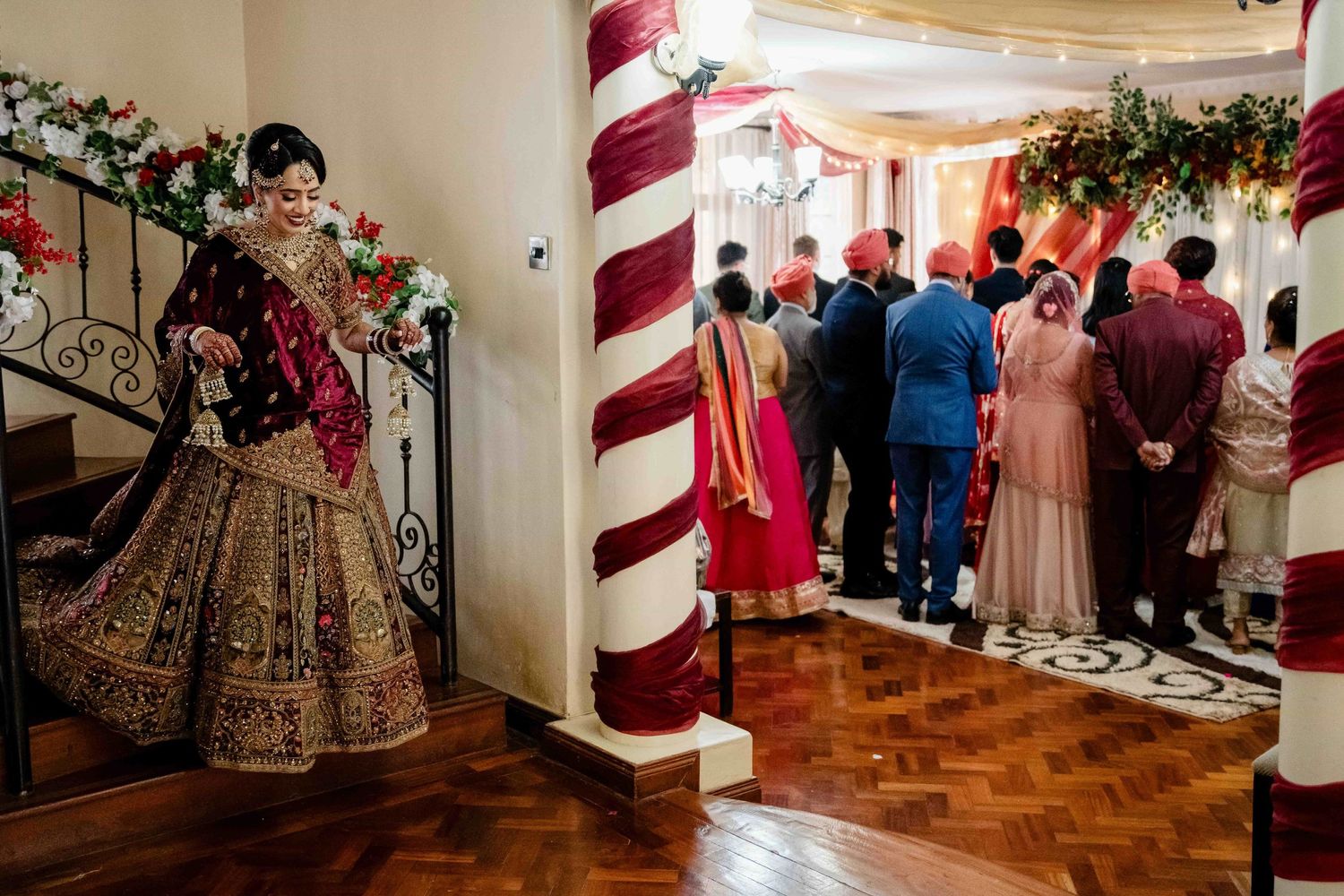 Wedding guests gather in a decorated hallway while a bride in elaborate lehenga poses near a staircase.