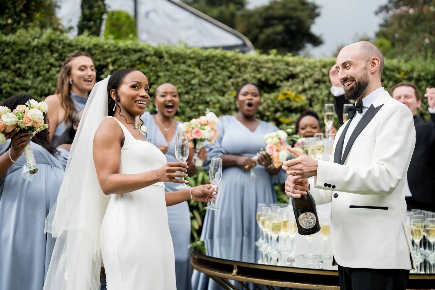 Wedding party guests in light blue dresses celebrate with champagne in an outdoor garden setting.