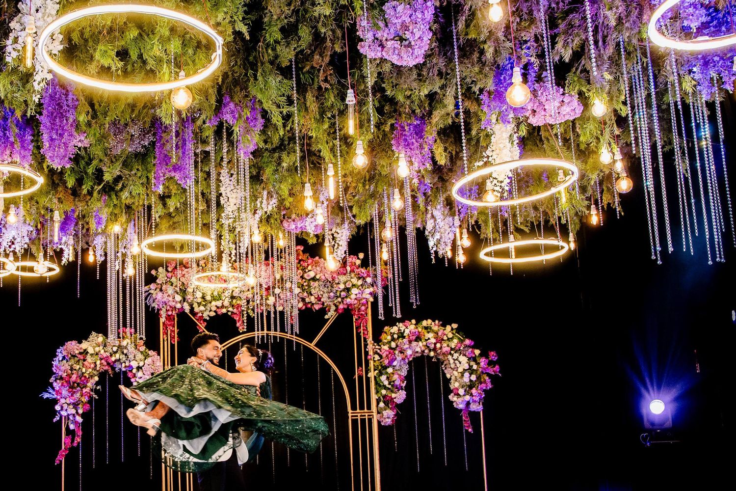 Elegant wedding reception decor with hanging crystal chandeliers and purple floral arrangements illuminated by golden lights.