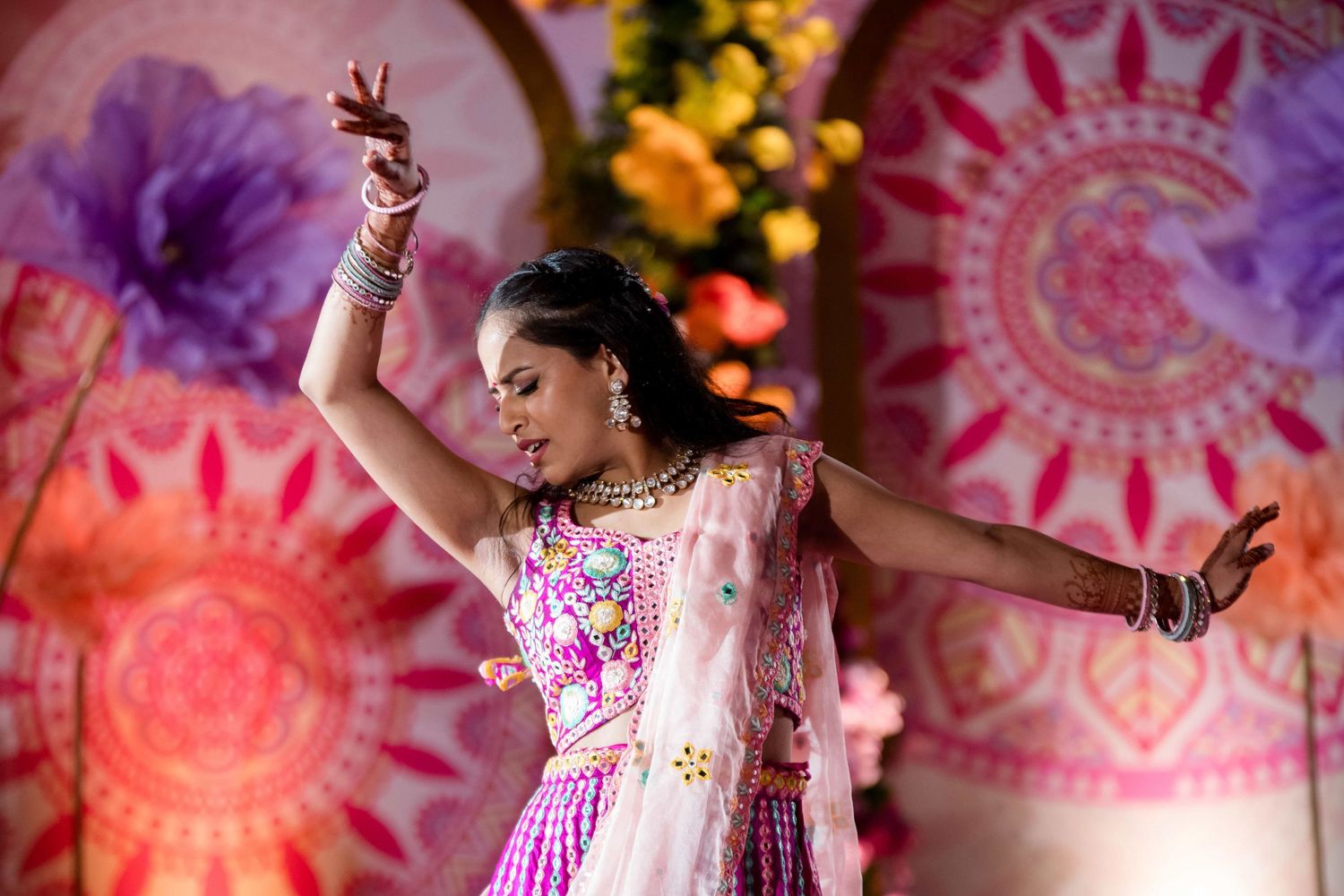 A dancer in a pink lehenga performs graceful movements against a colorful mandala backdrop.
