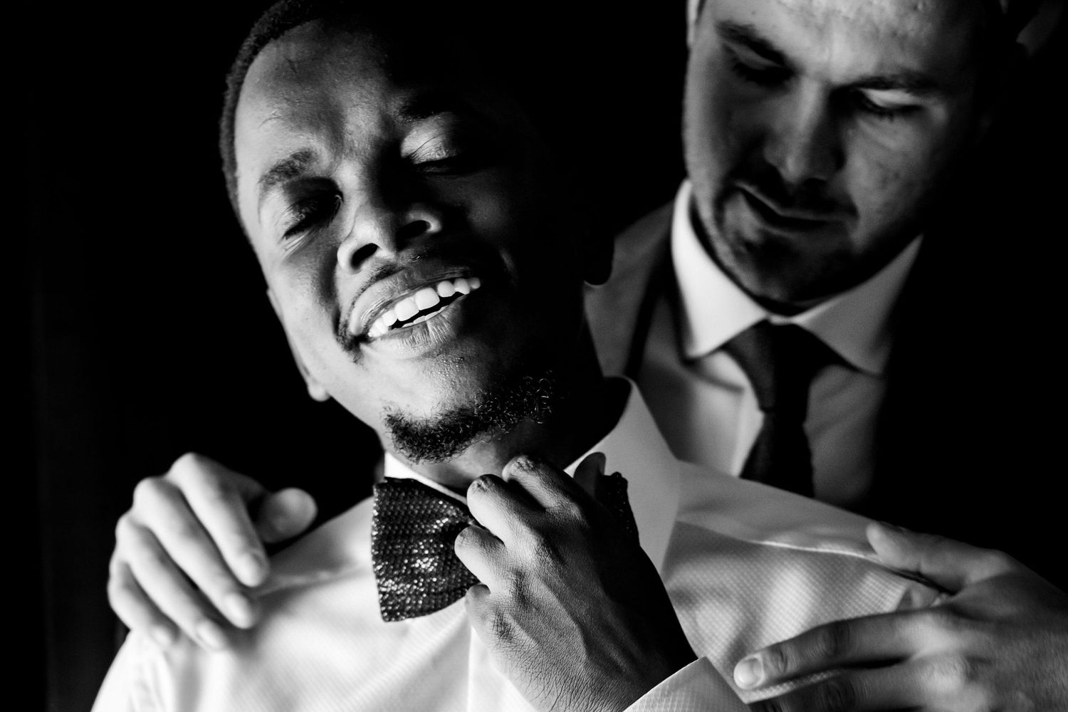 Black and white candid photo showing two men laughing while adjusting formal wear.