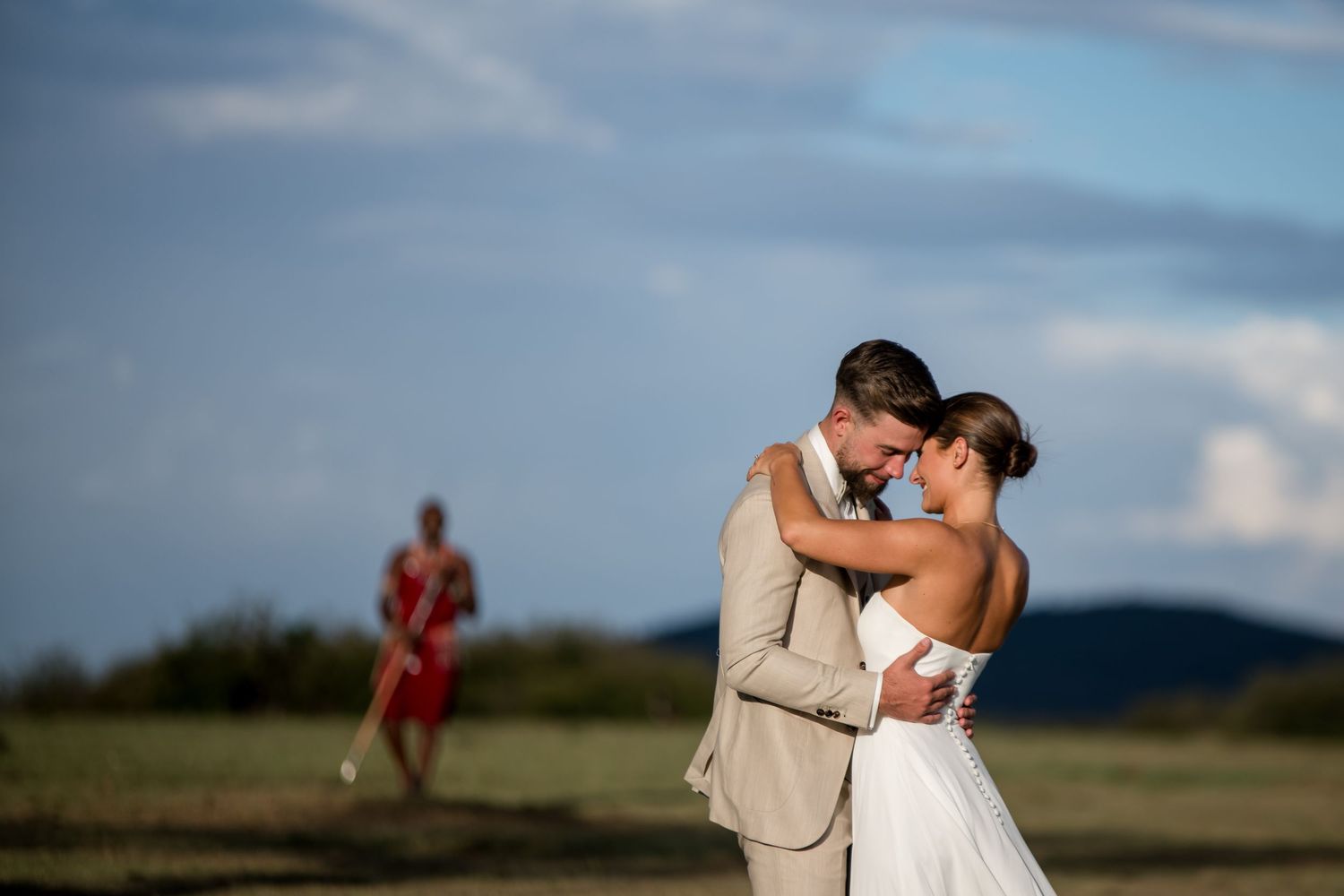 A bride and groom share a romantic embrace outdoors with a guest in red visible in the background.
