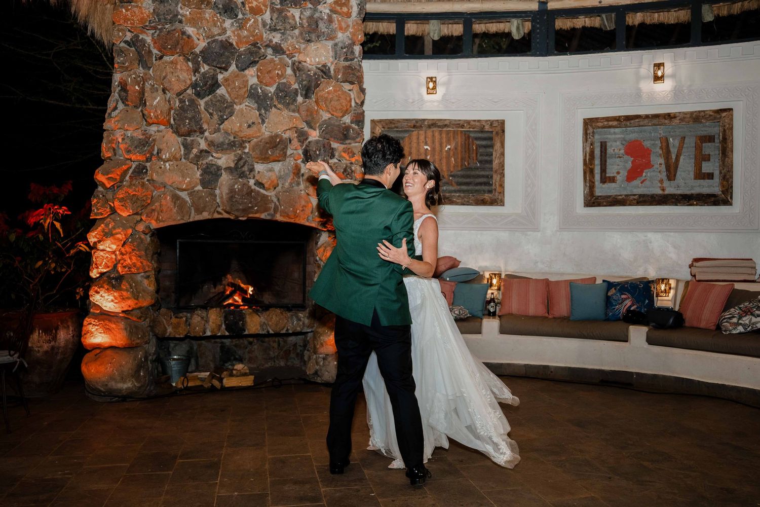 A couple shares their first dance in front of a rustic stone fireplace with a glowing LOVE sign on the wall.